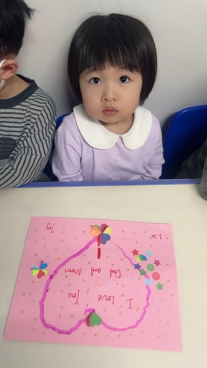 小女孩端坐桌前，展示亲手制作的I Love You Dad and Mom粉色爱心贺卡