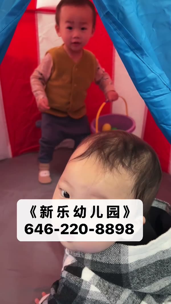 两个小朋友在红蓝色游戏帐篷里玩耍，其中一个提着装满彩色球的小桶